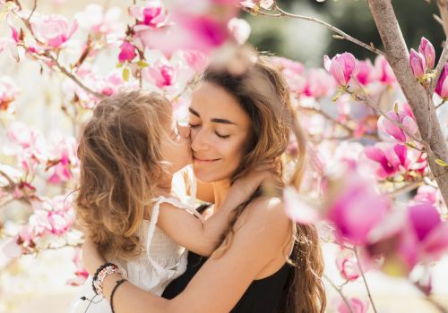 A woman and a young girl share an affectionate moment surrounded by pink flowers on a sunny day.