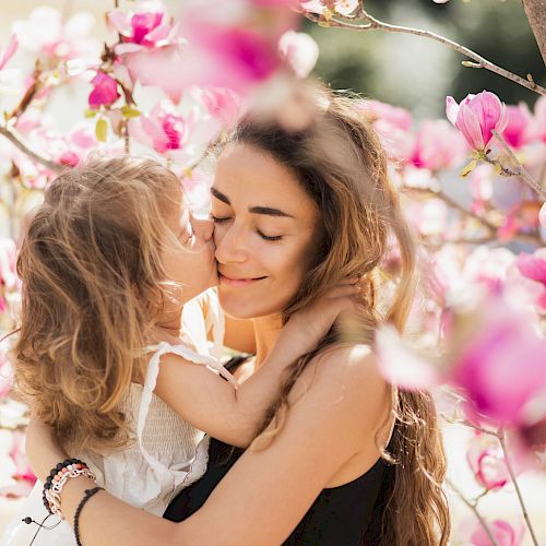 A woman and a young girl share an affectionate moment surrounded by pink flowers on a sunny day.