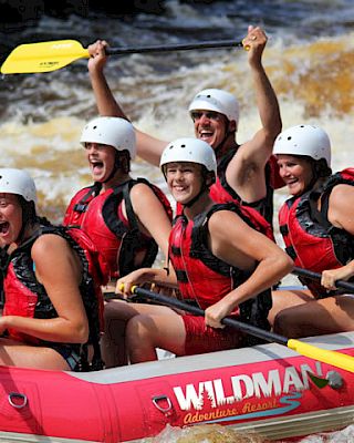 Six people in safety gear white water rafting in an inflatable boat, all wearing helmets and life jackets, holding paddles, and cheering excitedly.