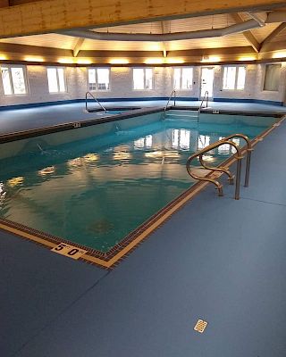 An indoor swimming pool with blue flooring. It has handrails, windows, and a wooden ceiling. The depth marker reads 