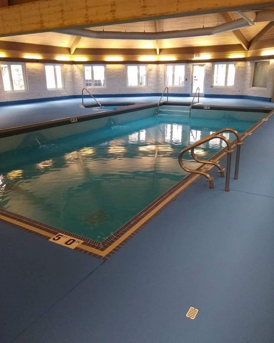 An indoor swimming pool with blue flooring. It has handrails, windows, and a wooden ceiling. The depth marker reads "5.0 ft" at one end.