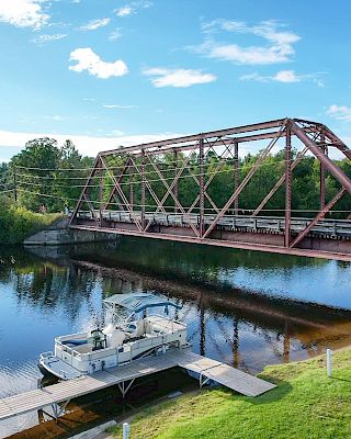 A red truss bridge spans over a calm river with a docked boat nearby, surrounded by trees and green grass on a sunny day.