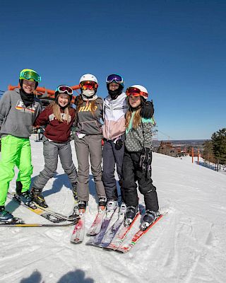 A group of five people wearing ski gear are posing on a snowy slope with clear blue skies in the background, enjoying their time.