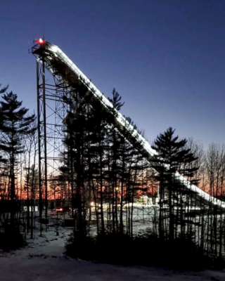 The image shows a large, illuminated ski jumping ramp surrounded by trees at dusk. The sky is dark with a hint of sunset colors.