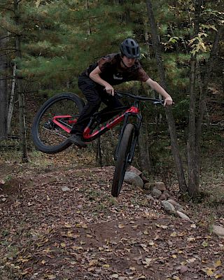 A person is performing a jump on a mountain bike in a forested trail area, wearing a helmet and protective gear.