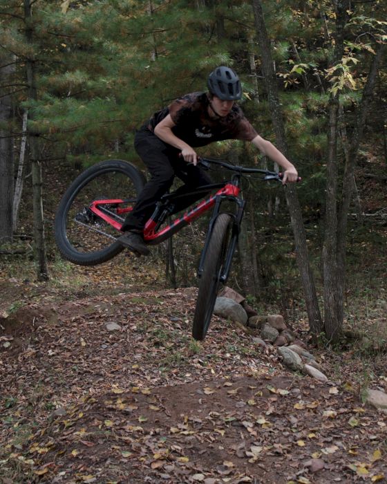 A person is performing a jump on a mountain bike in a forested trail area, wearing a helmet and protective gear.