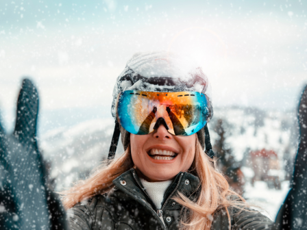 A person in winter gear and goggles smiles in a snowy outdoor setting, with arms raised.