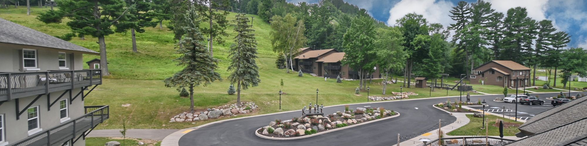 A scenic view of a hillside resort with buildings, trees, a curved road, and outdoor seating areas under a partly cloudy sky.