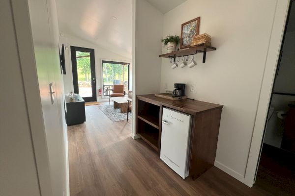 The image shows a small, modern kitchenette with a mini-fridge, open shelving, and a coffee maker. Living area with a couch visible in the background.