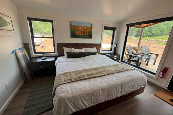 A cozy, modern bedroom with a large bed, two windows, and a glass door leading to a deck with chairs, overlooking a scenic landscape.