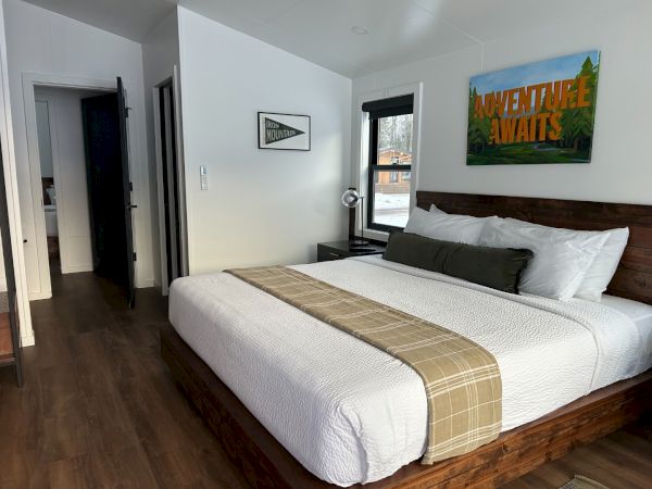 A cozy bedroom with a large bed, wooden headboard, and decor. "Adventure Awaits" sign and a window with a snowy view complete the scene.