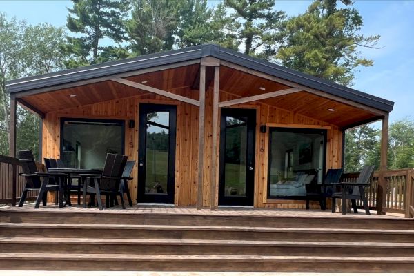 A wooden cabin with two glass doors, outdoor seating on a spacious deck, surrounded by trees and nature, with stairs leading up.