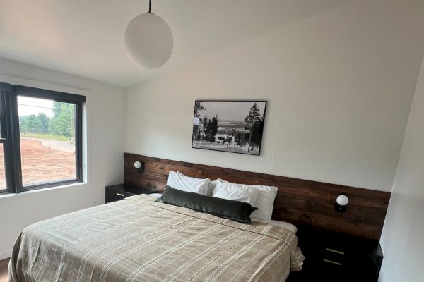 The image shows a modern bedroom with a large bed, nightstands, a hanging light fixture, and a window with a view of an outside area.