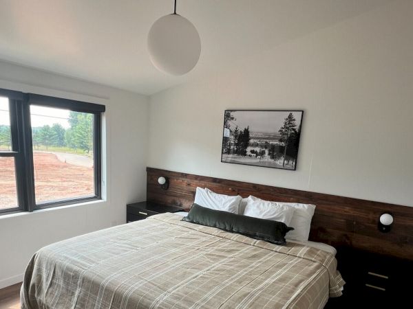 The image shows a modern bedroom with a large bed, nightstands, a hanging light fixture, and a window with a view of an outside area.