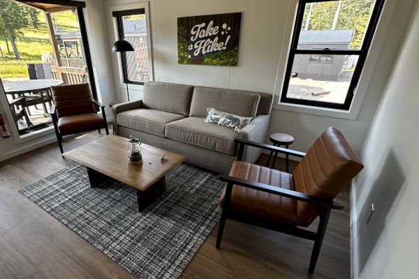 A cozy living room with a sofa, two chairs, coffee table, and a "Take a Hike!" sign on the wall, overlooking a porch and greenery outside.