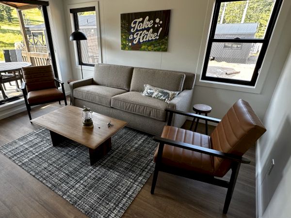 A cozy living room with a sofa, two chairs, coffee table, and a "Take a Hike!" sign on the wall, overlooking a porch and greenery outside.