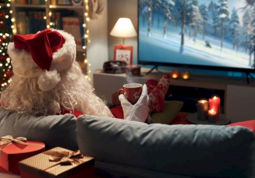 A person in a Santa outfit watches TV in a cozy, festive living room with wrapped gifts, candles, and a mug in hand.