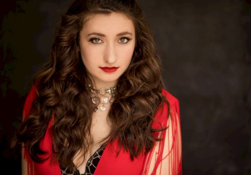 A woman with wavy brown hair, wearing a red outfit and a jeweled necklace, poses against a dark background.