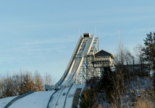 The image shows a snow-covered ski jump ramp with a structure at the top, surrounded by trees, under a clear blue sky.