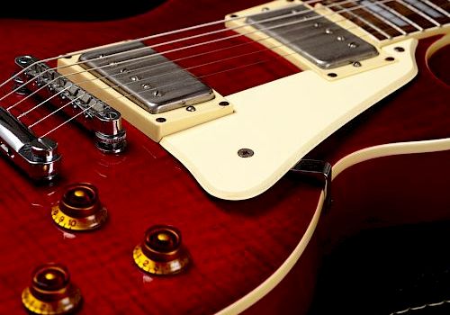 A close-up of a red electric guitar with a white pickguard and four control knobs is shown.