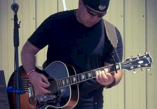 A person wearing a black cap and t-shirt is playing an acoustic guitar on an outdoor stage beside a microphone, with a background of a metallic wall.