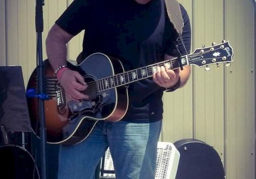 A person in a black shirt and hat is playing an acoustic guitar outdoors, standing near a microphone stand and some equipment.