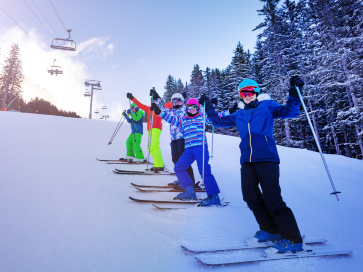 A group of people skiing on a snowy slope, wearing colorful winter gear and helmets, with trees and ski lifts in the background.