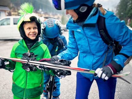 A group of people in ski gear are preparing for skiing; the child in green is holding skis, smiling, with others assisting.