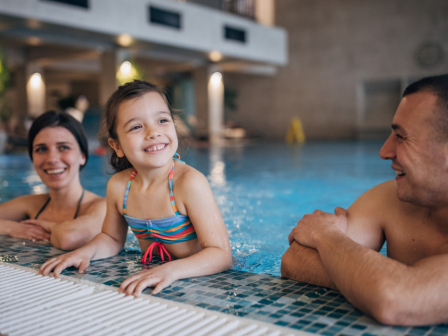 A child and two adults are enjoying time in an indoor swimming pool, smiling and resting at the pool's edge.