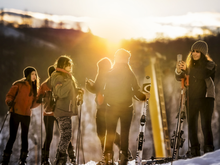 A group of people standing with skis, enjoying a snowy landscape at sunset, while one person takes a photo with a smartphone.