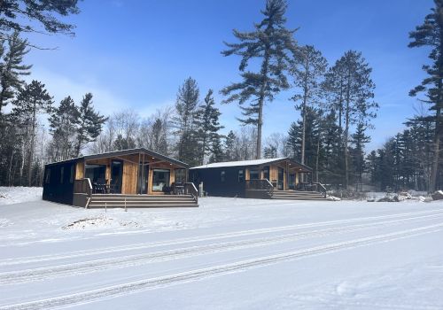 The image shows two wooden cabins surrounded by snow and pine trees under a clear blue sky.