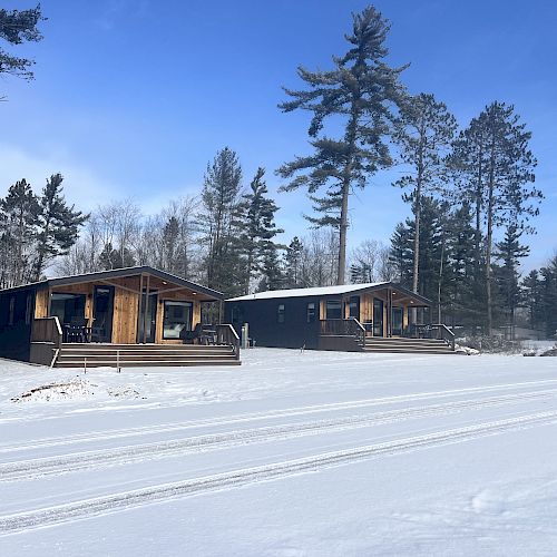 The image shows two wooden cabins surrounded by snow and pine trees under a clear blue sky.