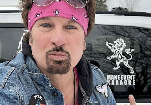 A person with a pink headband and sunglasses making a gesture in front of a vehicle with "Mane Event Karaoke" and a lion decal on the window.