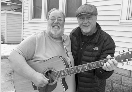 Two older men are smiling and posing with an acoustic guitar in front of a house, both looking happy.