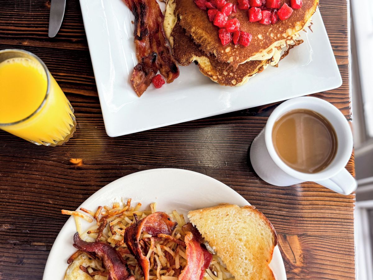 A breakfast spread with pancakes topped with strawberries, bacon, eggs, toast, hash browns, orange juice, and coffee.