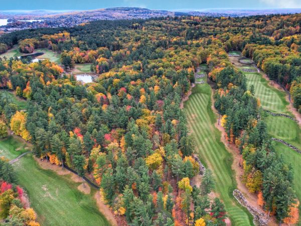 Aerial view of a golf course amidst a forest with fall foliage, showcasing green fairways, dense colorful trees, and a distant lake.