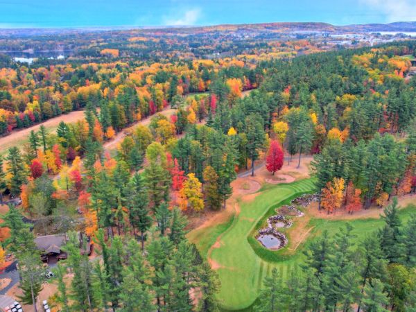 Aerial view of a golf course surrounded by a forest with vibrant autumn foliage in red, orange, and green hues.