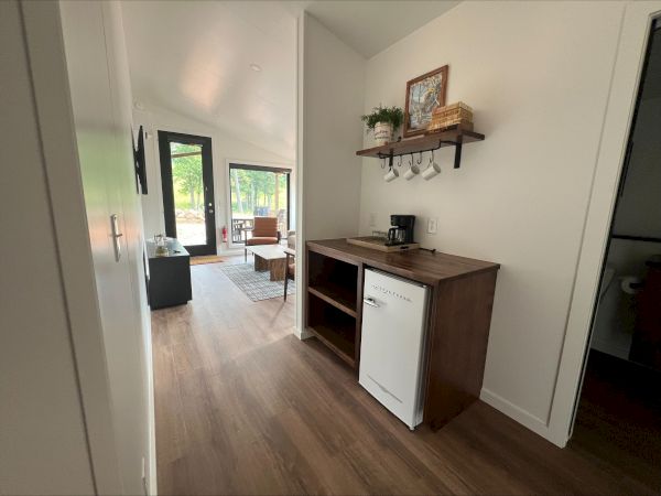 A bright room with wooden floor, a small fridge, coffee maker, shelf with decor, living area with chairs, and large window overlooking trees.
