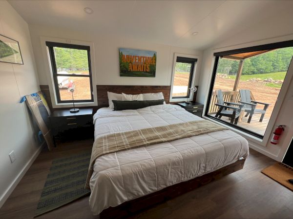 A cozy bedroom with a large bed, windows showing a scenic view, chairs outside, and a sign reading "Adventure Awaits."