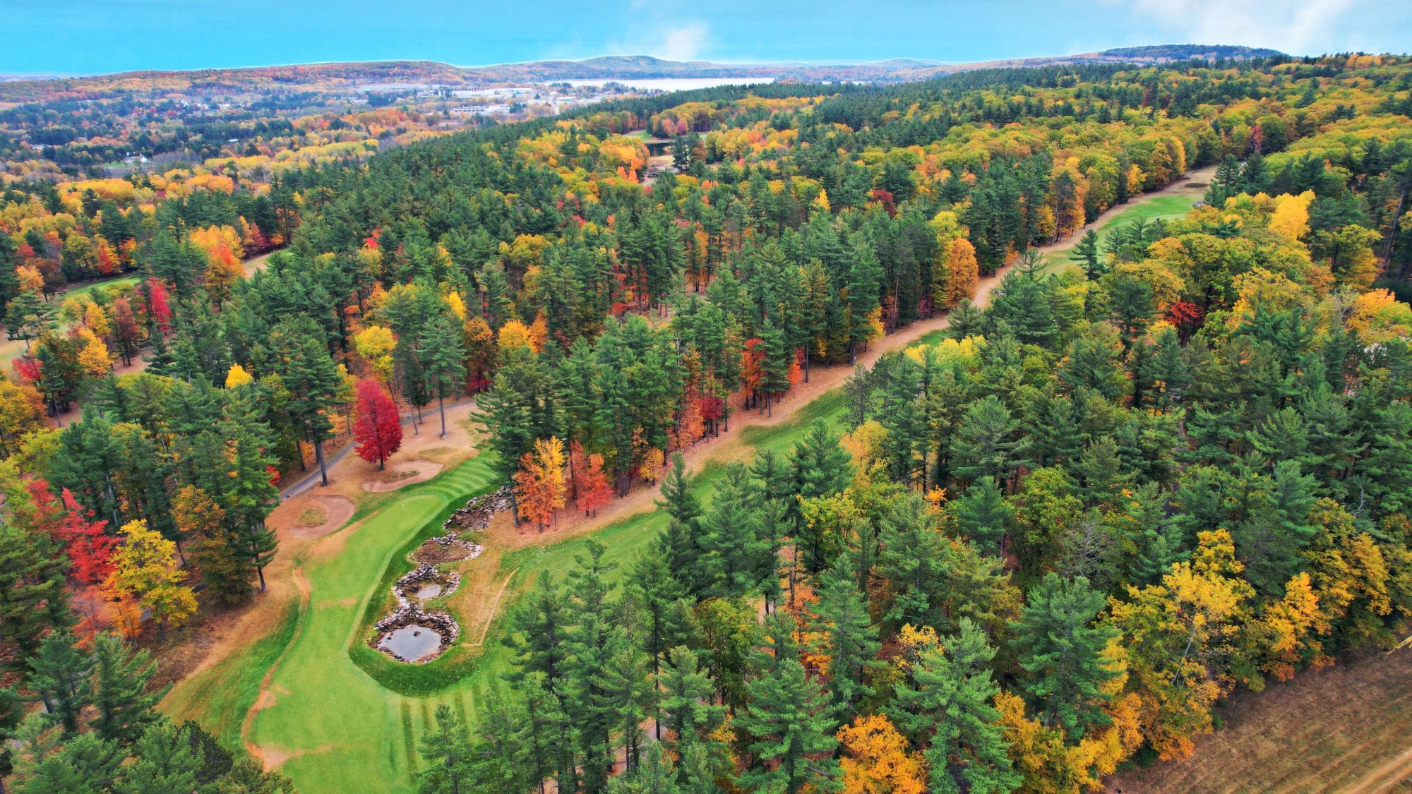 An aerial view shows a lush forest with vibrant autumn colors, winding paths, and a small pond surrounded by trees.