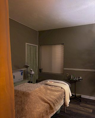 A cozy spa room with a massage table, soft blankets, and dim lighting. A small table with towels and bottles sits to the side.