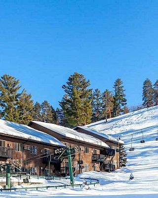 A snowy landscape features a ski resort with buildings, ski lifts, and surrounding evergreen trees under a clear blue sky.