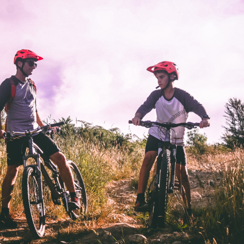 Two people wearing helmets are on mountain bikes, paused on a dirt trail, surrounded by tall grass and trees under a cloudy sky.