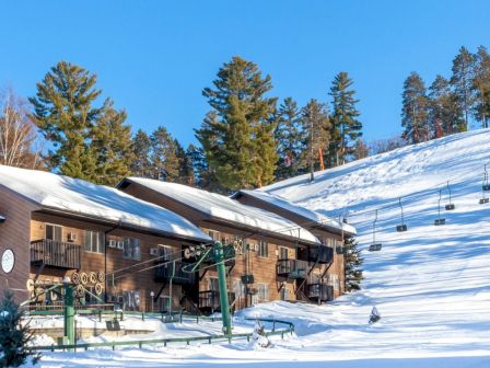 A snowy ski resort with wooden buildings, ski lifts, and pine trees visible on the hill in the background.