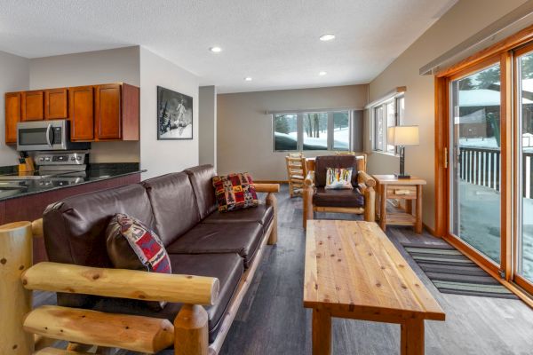 A cozy living room with a brown couch, chairs, wooden table, and rugs, adjacent to a kitchen with wooden cabinets and stainless appliances.