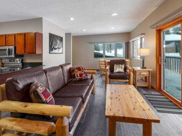 A cozy living room with a brown couch, chairs, wooden table, and rugs, adjacent to a kitchen with wooden cabinets and stainless appliances.