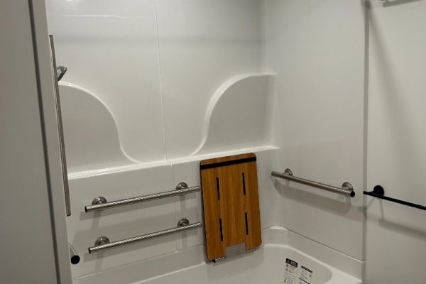 A bathroom features a bathtub with grab bars, a fold-down wooden seat, a shower rod, and overhead lighting.