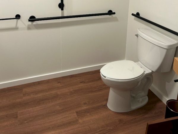A bathroom with a toilet, wooden floor, and multiple black grab bars on white walls for accessibility.