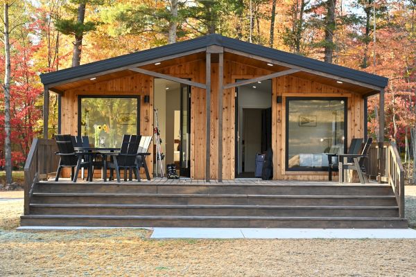 A modern cabin with large windows, a wooden patio, and outdoor furniture is set amid autumn trees with colorful leaves.