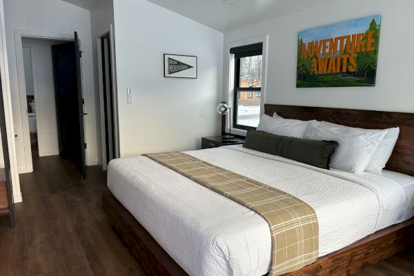 The image shows a cozy bedroom with a large bed, a plaid runner, wall art saying "Adventure Awaits," and a small lamp by the window.